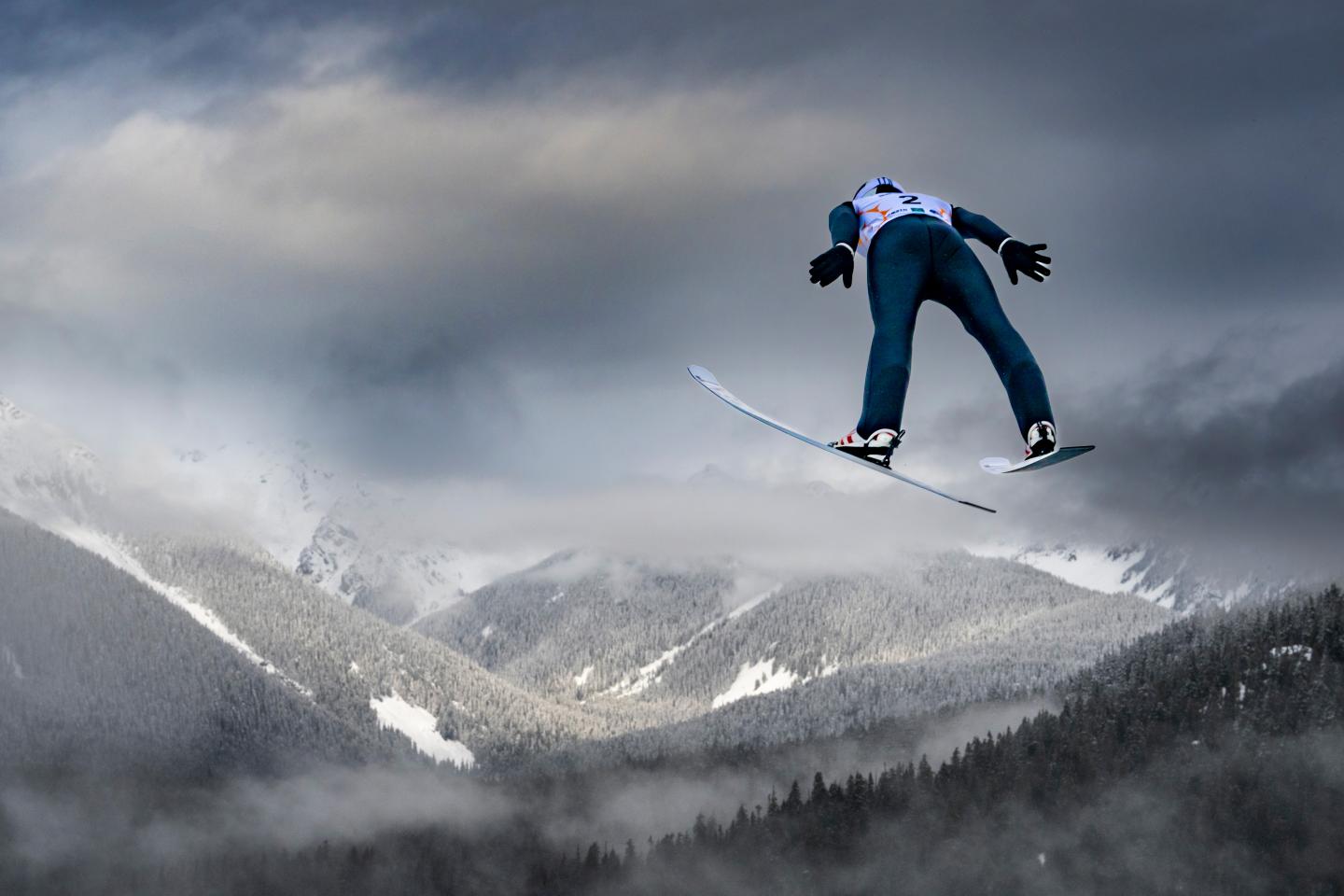 nordic skier after a jump, in the air, with cloudy mountains in the background