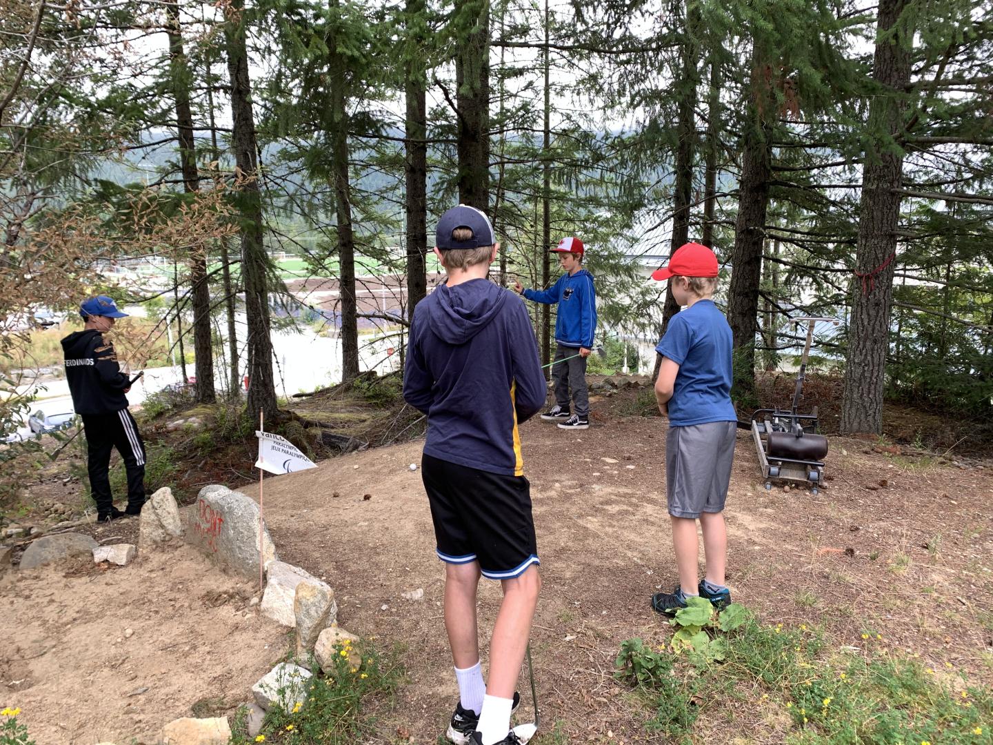 Four people stand in a forest clearing, one gesturing towards the trees.