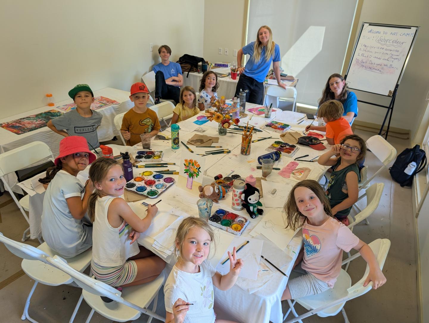 Children seated around a table, painting and smiling, with an instructor standing nearby.